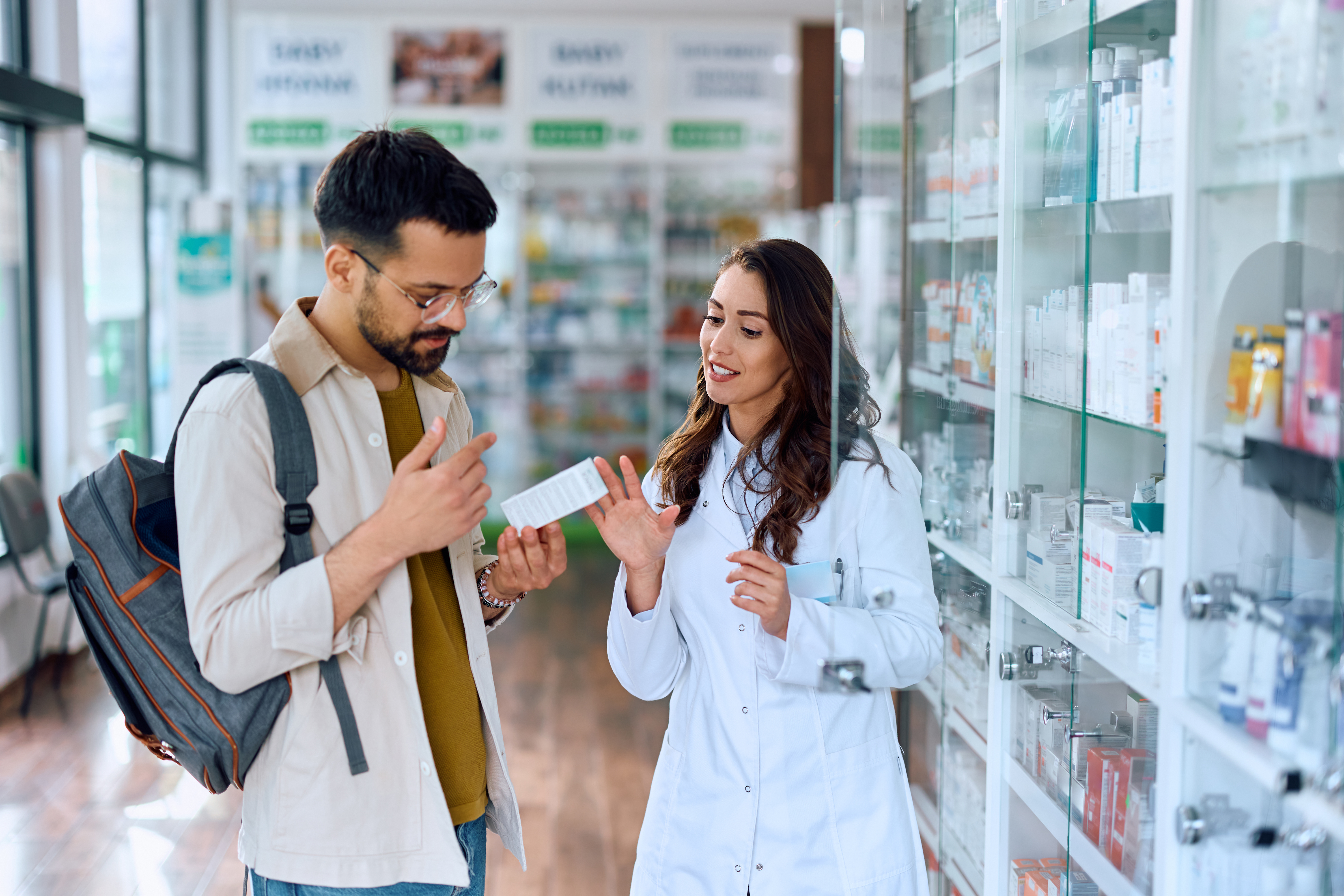 Female pharmacist standing beside glass shelving showing a customer a prescription